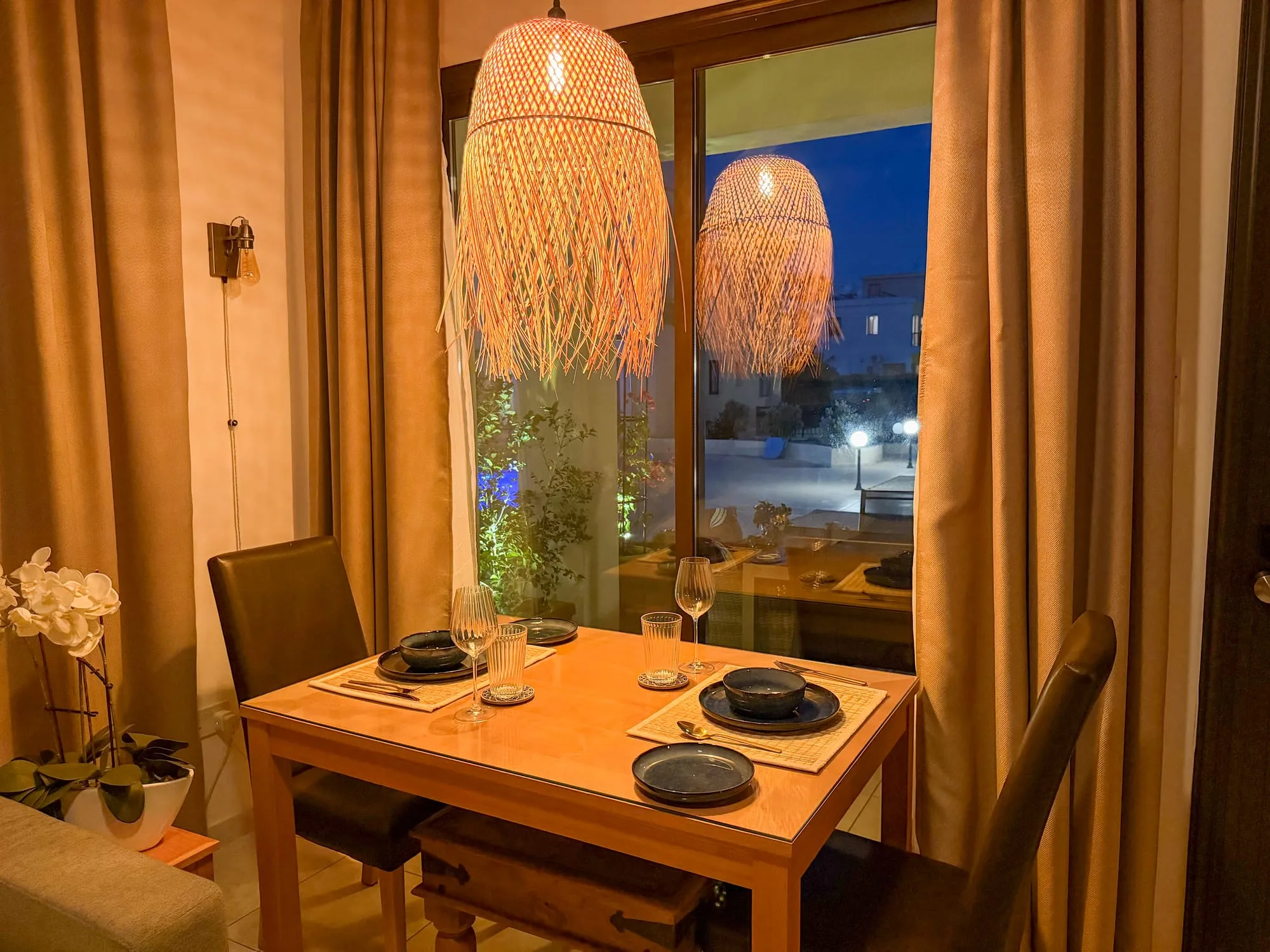The dining table set for two in the evening, under woven pendant lights by the window